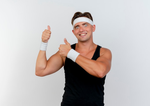 Glimlachende jonge knappe sportieve mens die hoofdband en polsbandjes draagt die duimen tonen die omhoog op witte muur worden geïsoleerd