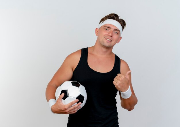 Glimlachende jonge knappe sportieve mens die hoofdband en polsbandjes draagt die voetbal houden en duim tonen die op witte muur wordt geïsoleerd