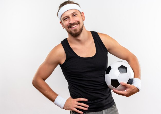 Glimlachende jonge knappe sportieve mens die hoofdband en polsbandjes draagt die voetbal met hand op taille houden die op witte ruimte wordt geïsoleerd