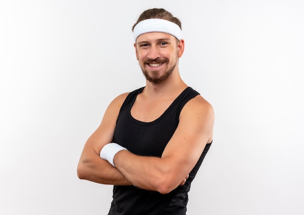 Glimlachende jonge knappe sportieve mens die hoofdband en polsbandjes draagt die zich met gesloten houding bevinden die op witte ruimte wordt geïsoleerd