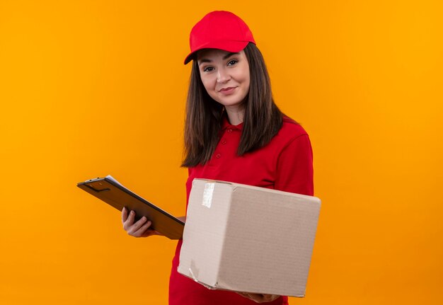 Glimlachende jonge leveringsvrouw die rode t-shirt in rode GLB draagt die een doos en een klembord op geïsoleerde gele muur houdt