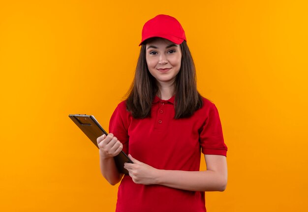 Glimlachende jonge leveringsvrouw die rode t-shirt in rode GLB draagt die een klembord op geïsoleerde gele muur houdt