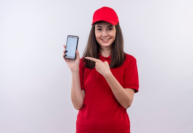 Glimlachende jonge leveringsvrouw die rode t-shirt in rode GLB draagt die een telefoon op geïsoleerde witte muur houdt