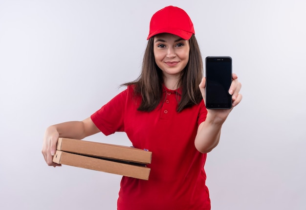 Glimlachende jonge leveringsvrouw die rode t-shirt in rode glb dragen die pizzadoos en telefoon op geïsoleerde witte muur houden