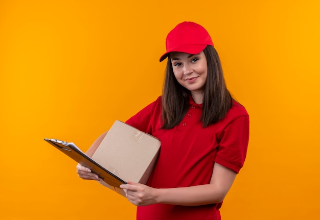 Glimlachende jonge leveringsvrouw die rode t-shirt in rode pet draagt die een doos en flipboard op geïsoleerde gele muur houdt