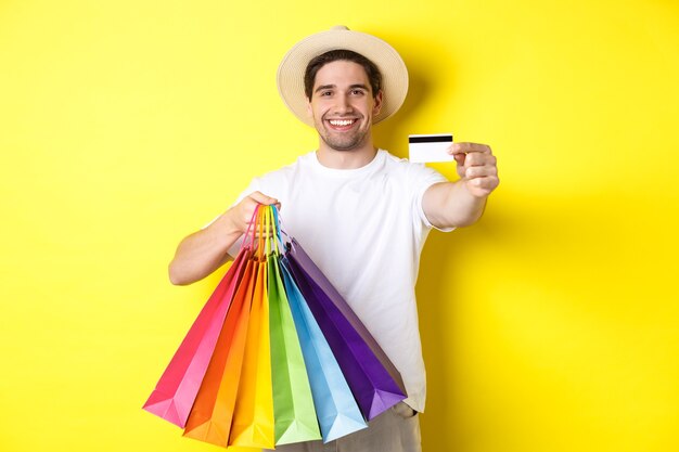 Glimlachende jonge man die dingen koopt met een creditcard, boodschappentassen vasthoudt en er gelukkig uitziet, staande over een gele achtergrond.
