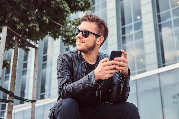 Glimlachende jonge man in zonnebril met stijlvol haar gekleed in zwart leren jas houdt smartphone vast terwijl hij in de buurt van wolkenkrabber zit.