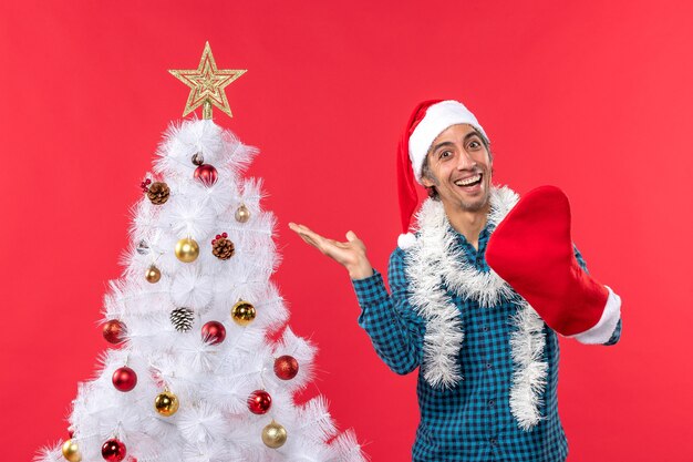 Glimlachende jonge man met kerstman hoed in een blauw gestript shirt en het dragen van zijn kerstsok in de buurt van kerstboom op rode beelden