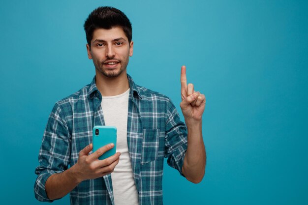 Glimlachende jonge man met mobiele telefoon kijkend naar camera die omhoog wijst geïsoleerd op blauwe achtergrond met kopieerruimte