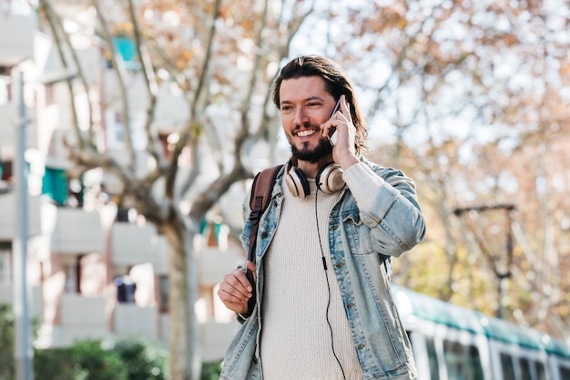 Glimlachende jonge man met zijn rugzak praten op mobiele telefoon in de open lucht