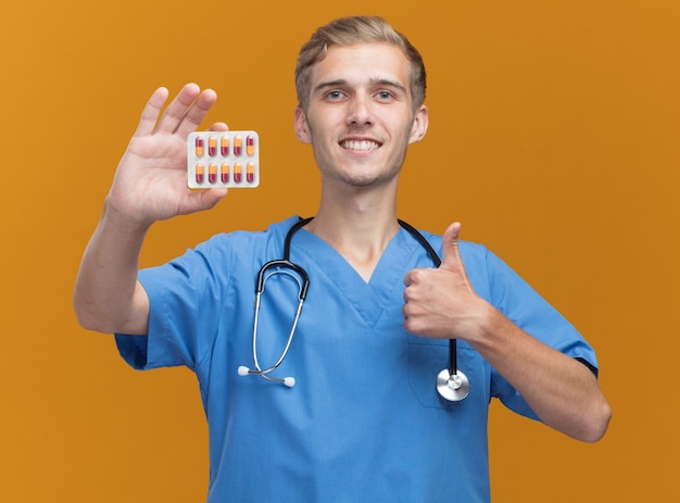 Glimlachende jonge mannelijke arts die arts eenvormig met de pillen van de stethoscoopholding draagt die duim tonen die omhoog op oranje muur wordt geïsoleerd