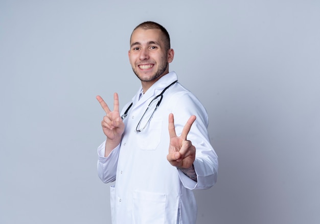 Glimlachende jonge mannelijke arts die medisch kleed en een stethoscoop om zijn hals draagt die zich in profielmening bevindt die vredestekens doet die op witte muur worden geïsoleerd