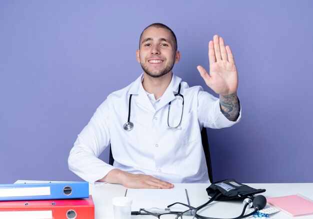 Glimlachende jonge mannelijke arts die medische mantel en stethoscoop draagt ?? die aan bureau zit met uitrustingsstukken die hand op bureau zetten en gebaren stoppen aan voorzijde geïsoleerd op paarse muur