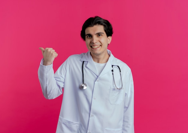 Glimlachende jonge mannelijke arts die medische mantel en stethoscoop draagt die achter geïsoleerd op roze muur met exemplaarruimte richten