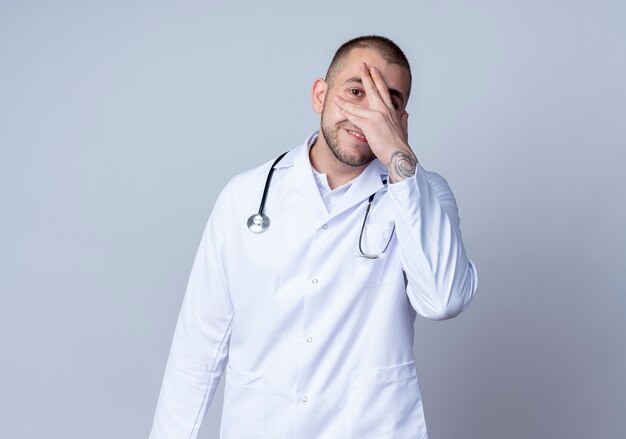 Glimlachende jonge mannelijke arts die medische mantel en stethoscoop draagt die hand op gezicht zet en naar voorzijde kijkt door vingers om zijn hals die op witte muur wordt geïsoleerd