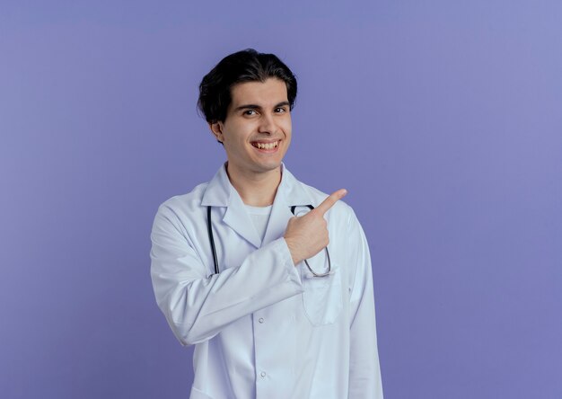 Glimlachende jonge mannelijke arts die medische mantel en stethoscoop draagt die naar kant richt die op purpere muur met exemplaarruimte wordt geïsoleerd