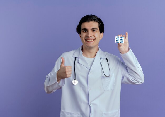 Glimlachende jonge mannelijke arts die medische mantel en stethoscoop draagt die pak capsules en duim toont die omhoog op purpere muur met exemplaarruimte worden geïsoleerd