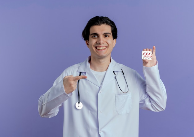 Glimlachende jonge mannelijke arts die medische mantel en stethoscoop draagt die pak medische capsules toont die op het richten geïsoleerd op purpere muur