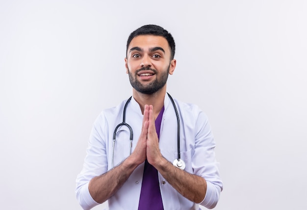 Glimlachende jonge mannelijke arts die stethoscoop medische toga draagt die bidgebaar op geïsoleerde witte achtergrond toont