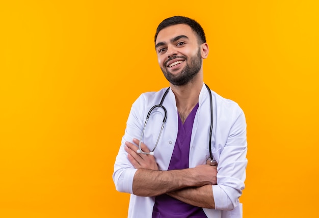 Glimlachende jonge mannelijke arts die stethoscoop medische toga draagt die handen op geïsoleerde gele achtergrond kruisen