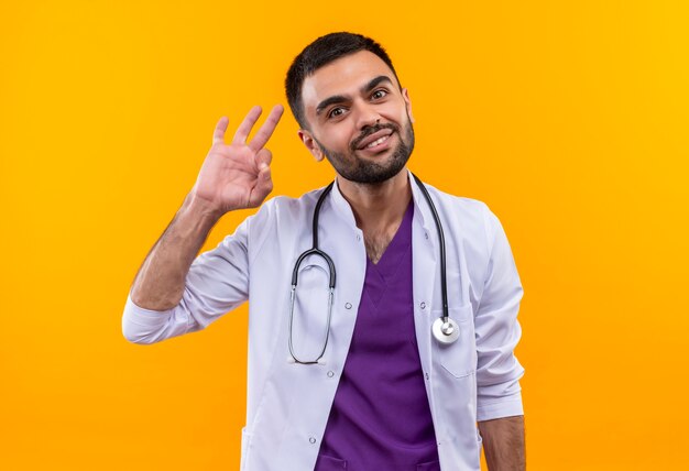 Glimlachende jonge mannelijke arts die stethoscoop medische toga draagt die okgebaar op geïsoleerde gele achtergrond toont