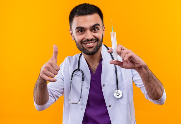 Glimlachende jonge mannelijke arts die stethoscoop medische toga draagt die spuit zijn duim tegen geïsoleerde gele achtergrond houdt