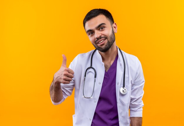 Glimlachende jonge mannelijke arts die stethoscoop medische toga draagt zijn duim omhoog op geïsoleerde gele achtergrond