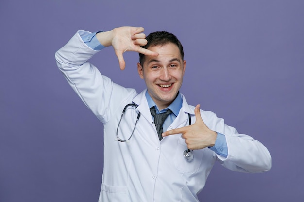 Glimlachende jonge mannelijke arts met een medisch gewaad en een stethoscoop om de nek die naar de camera kijkt en een framegebaar maakt dat op een paarse achtergrond wordt geïsoleerd