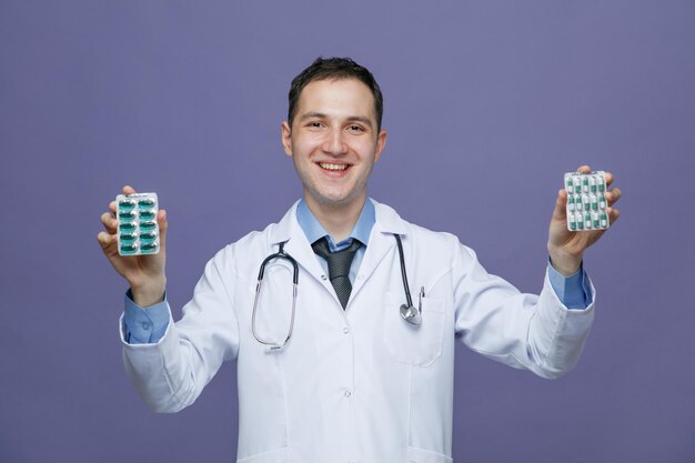 Glimlachende jonge mannelijke arts met een medisch gewaad en een stethoscoop om de nek, kijkend naar de camera met pakjes pillen geïsoleerd op een paarse achtergrond