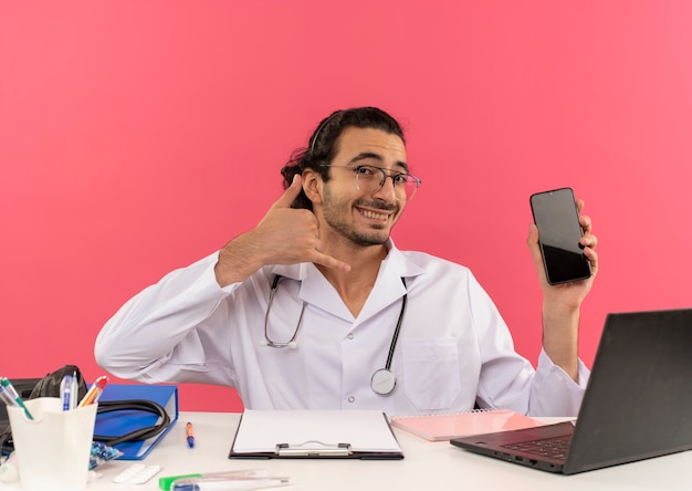 Glimlachende jonge mannelijke arts met een medische bril die een medische mantel draagt met een stethoscoop die aan het bureau zit