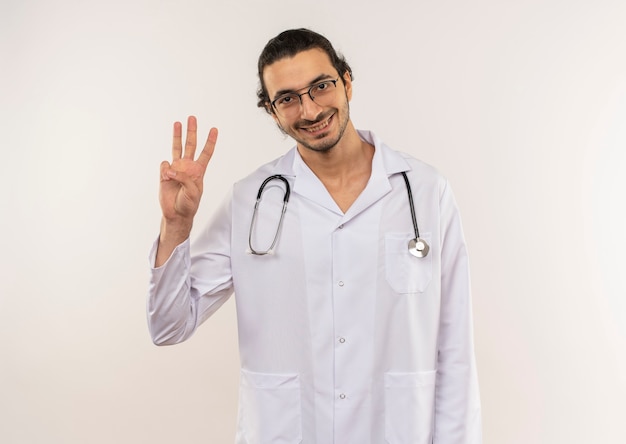 Glimlachende jonge mannelijke arts met optische bril die wit gewaad met stethoscoop draagt die drie op geïsoleerde witte muur met exemplaarruimte toont