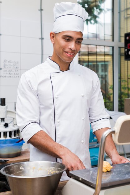 Glimlachende jonge mannelijke bakker die kneed het deeg op schaal in de commerciële keuken wegen