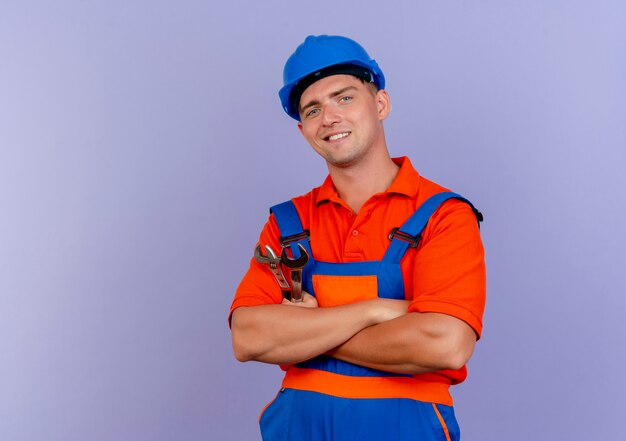 Glimlachende jonge mannelijke bouwer die eenvormig en veiligheidshelm draagt die handen op purple kruisen