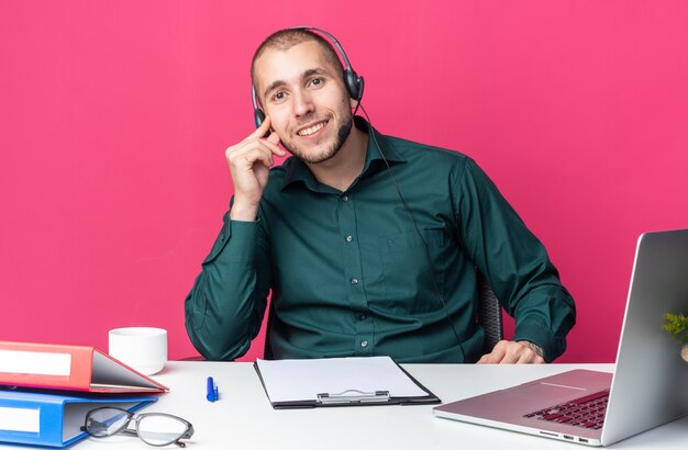 Glimlachende jonge mannelijke callcentermedewerker die een headset draagt die aan een bureau zit met kantoorhulpmiddelen