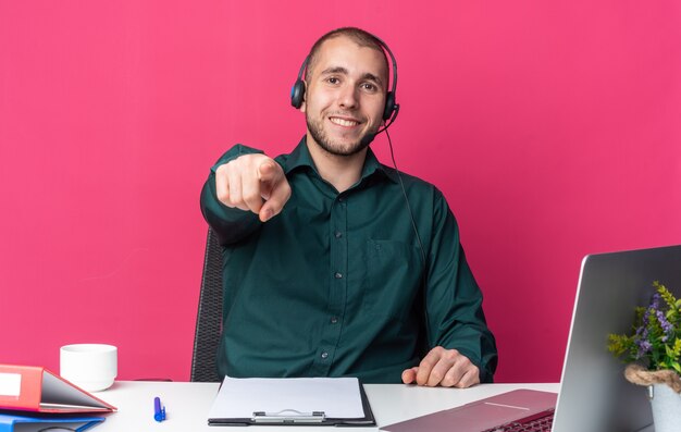 Glimlachende jonge mannelijke callcentermedewerker met een headset die aan het bureau zit met kantoorhulpmiddelen die je een gebaar laten zien