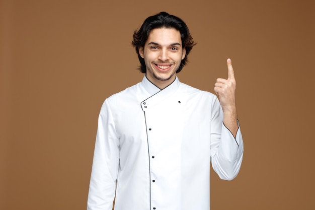 Glimlachende jonge mannelijke chef-kok die uniform draagt en naar de camera kijkt die omhoog wijst geïsoleerd op bruine achtergrond