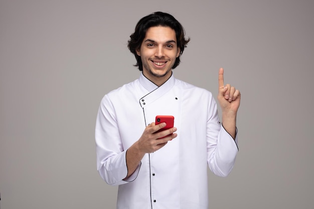 Glimlachende jonge mannelijke chef-kok in uniform met mobiele telefoon die naar de camera kijkt en omhoog wijst op een witte achtergrond Premium Foto