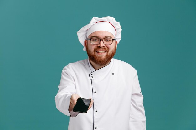 Glimlachende jonge mannelijke chef-kok met een uniforme bril en pet kijkend naar camera die mobiele telefoon uitrekt naar camera geïsoleerd op blauwe achtergrond