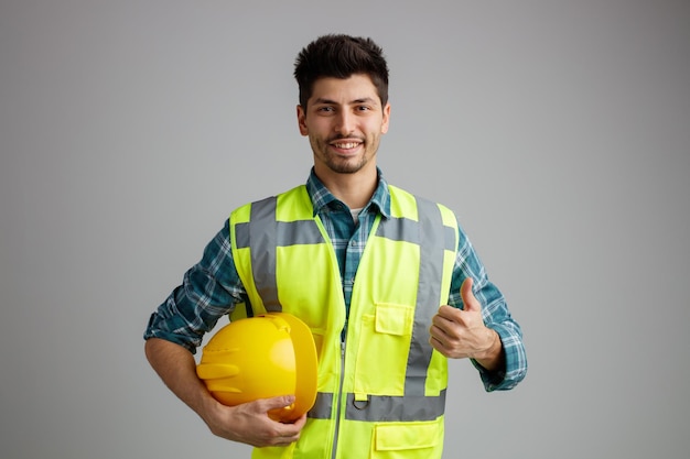 Glimlachende jonge mannelijke ingenieur die een uniforme veiligheidshelm draagt en naar de camera kijkt die duim omhoog laat zien op een witte achtergrond