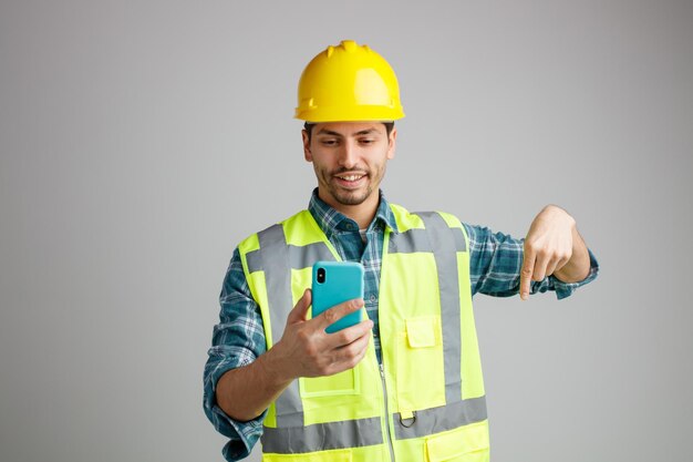 Glimlachende jonge mannelijke ingenieur die een veiligheidshelm en uniform draagt en naar een mobiele telefoon kijkt die naar beneden wijst, geïsoleerd op een witte achtergrond