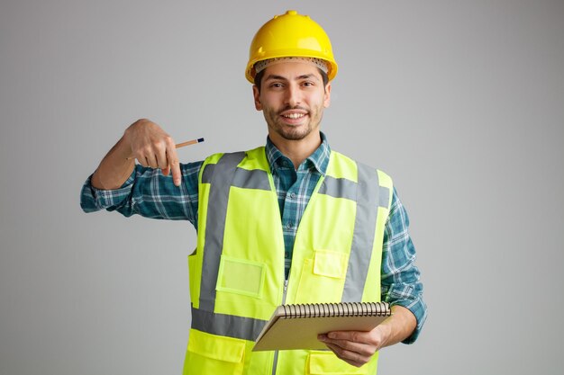 Glimlachende jonge mannelijke ingenieur met veiligheidshelm en uniform met notitieblok en potlood kijkend naar camera die naar beneden wijst, geïsoleerd op een witte achtergrond