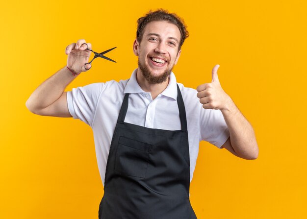 Glimlachende jonge mannelijke kapper die een uniforme holdingsschaar draagt die duim toont die op gele achtergrond wordt geïsoleerd