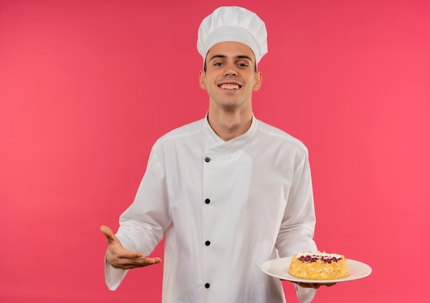Glimlachende jonge mannelijke kok die chef-kok eenvormige holding en punten met handcake op plaat draagt
