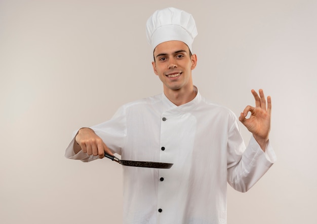Glimlachende jonge mannelijke kok die de pan van de chef-kok de eenvormige holding draagt die ok gebaar toont