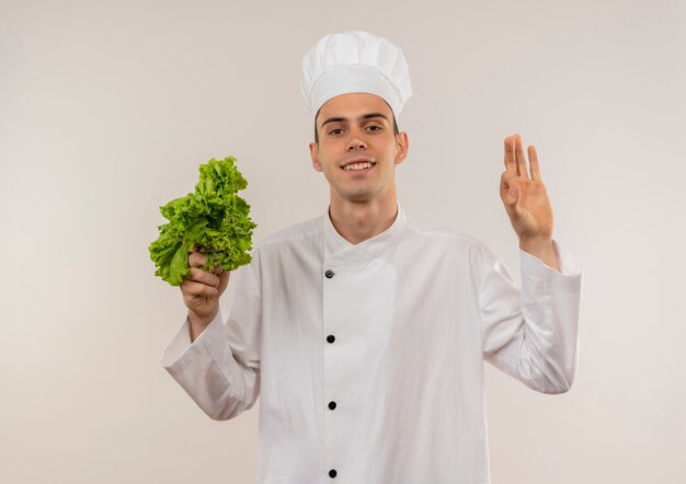 Glimlachende jonge mannelijke kok die de salade van de chef-kok eenvormige holding draagt die okgebaar toont