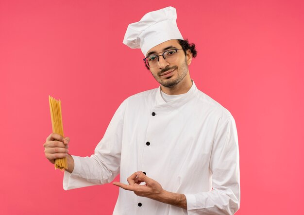 Glimlachende jonge mannelijke kok die eenvormige chef-kok en glazen houden en wijst naar spaghetti die op roze muur wordt geïsoleerd