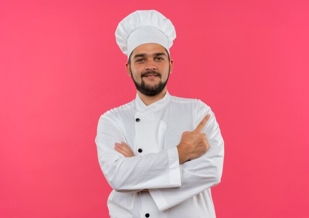 Glimlachende jonge mannelijke kok in eenvormige chef-kok die zich met gesloten houding bevindt en naar kant richt die op roze ruimte wordt geïsoleerd