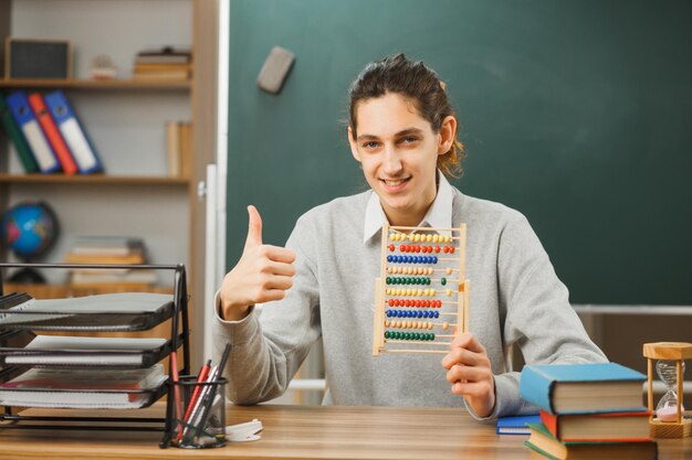 glimlachende jonge mannelijke leraar die aan het bureau zit en een telraam vasthoudt met duimen omhoog met schoolhulpmiddelen in de klas