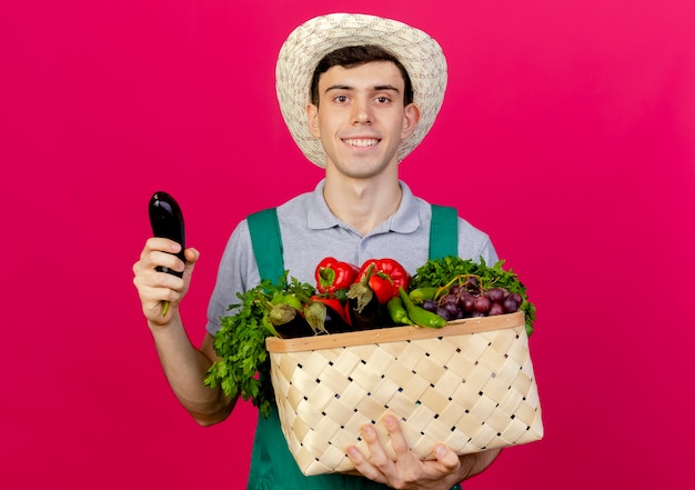 Glimlachende jonge mannelijke tuinman die een tuinhoed draagt, houdt een groentemand en aubergine vast