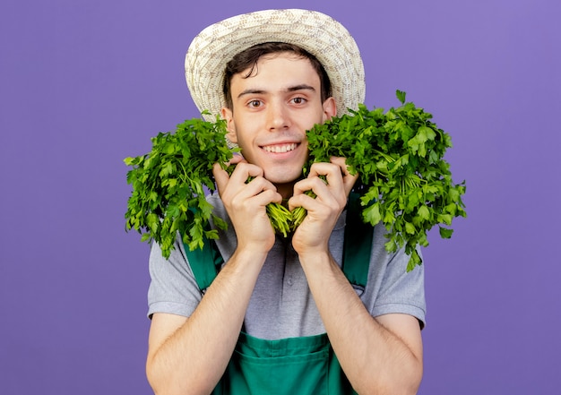 Glimlachende jonge mannelijke tuinman die een tuinhoed draagt, houdt koriander vast